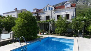 En swimmingpool med en metalstige, omgivet af frodig vegetation og en stor bygning med røde tegltage i baggrunden.