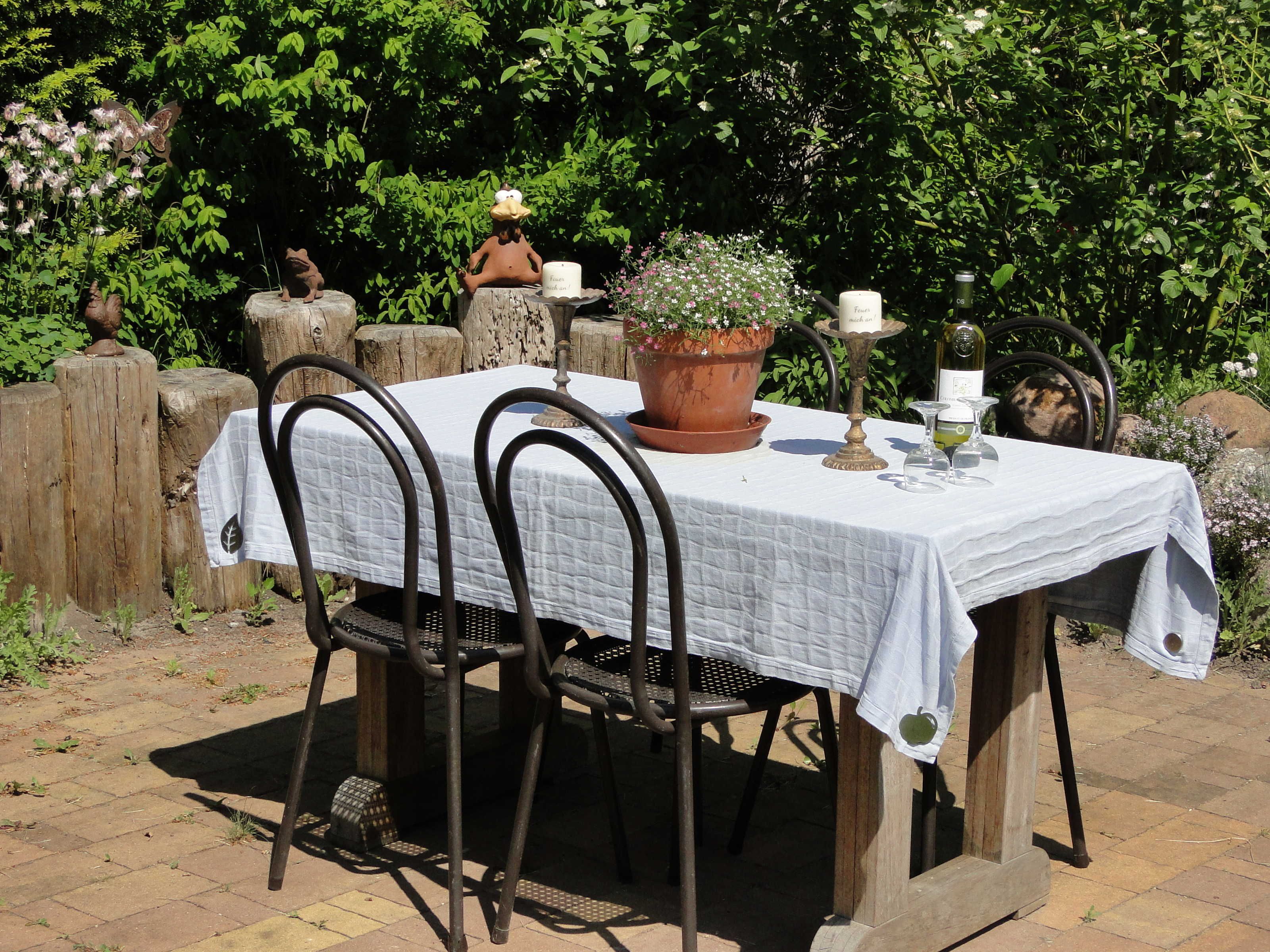Una mesa al aire libre con mantel blanco, sillas metálicas, una planta en maceta, una copa de vino y un candelabro.