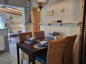 A dining area with wicker chairs, a dark table, and a blue cushion. There are wall-mounted shelves with decorative items and a hanging light fixture. - Blue Water 1 - Blue Water 1 (Wustrow)