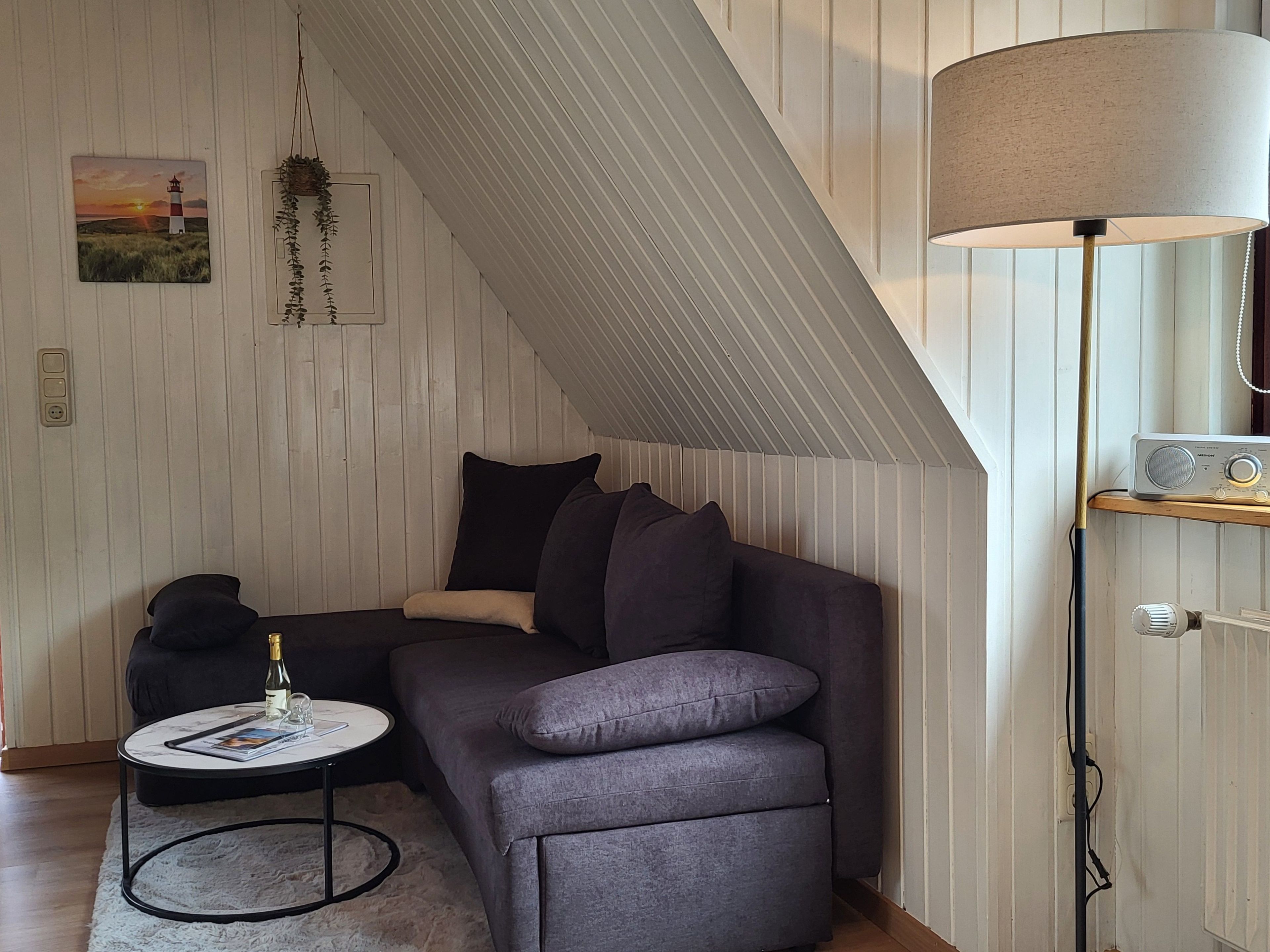 Ein Loft-Zimmer mit schrägem Dach, einem grauen Sofa, einem kleinen runden Tisch mit einer Flasche, einer Lampe und einem Radio.