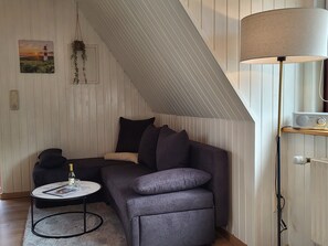 Een loft-achtige kamer met een schuine kap, een grijze bank, een klein rond tafeltje met een fles, een lamp en een radio.