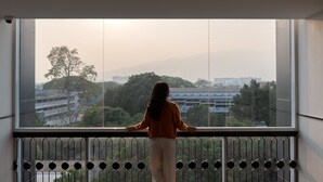 Eine Person steht an einem großen Fenster mit Blick auf eine Stadtlandschaft aus Gebäuden und Bäumen.