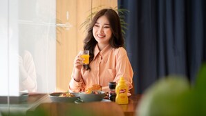 Eine Person sitzt an einem Tisch mit einem Glas Orangensaft, einer Schüssel Essen und einer gelben Flasche. Im Hintergrund steht eine Pflanze und ein Fenster bietet Blick auf Grünflächen.