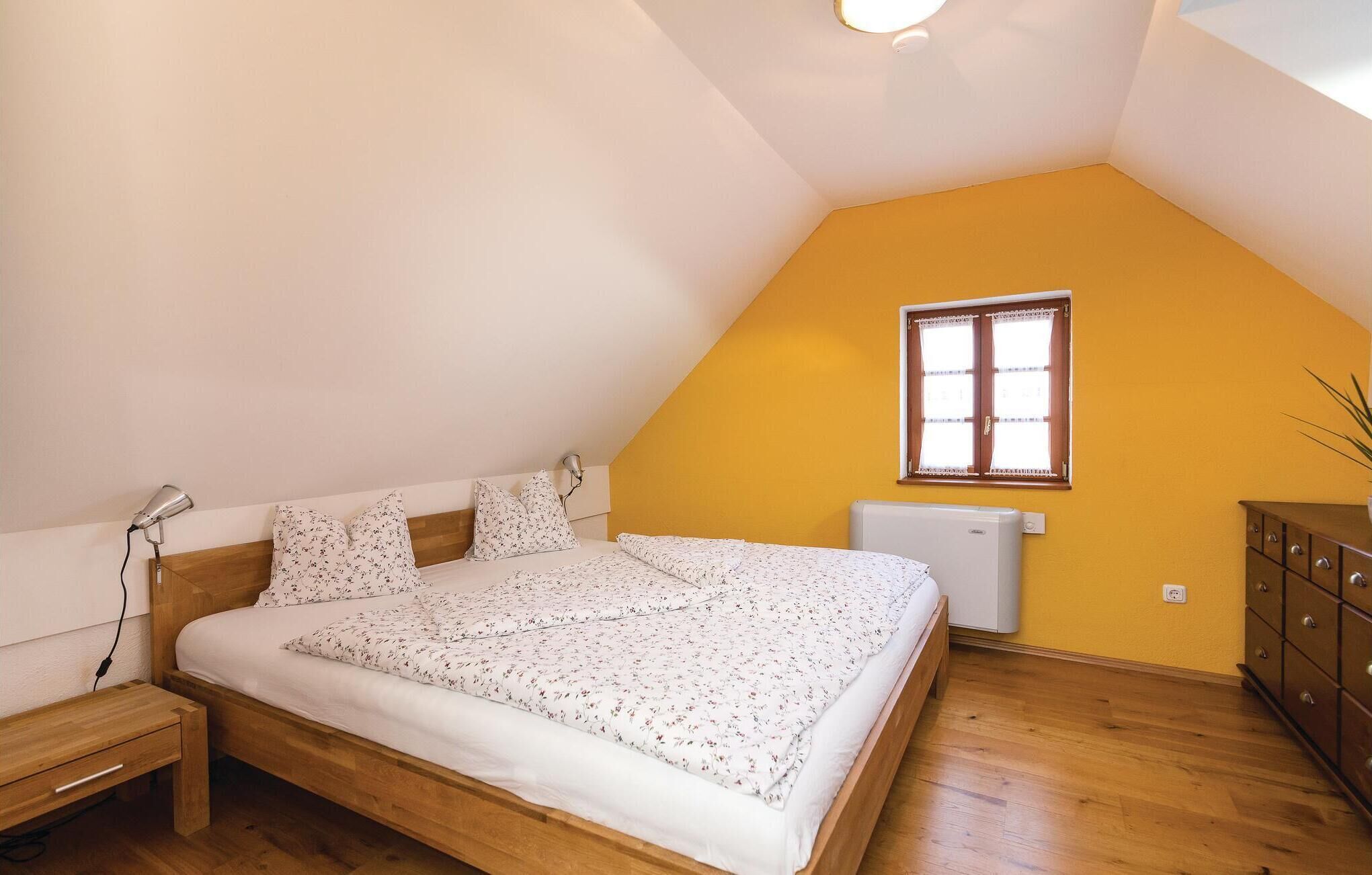 Un dormitorio con techo inclinado, una cama con ropa de cama floral, una lámpara de noche, un radiador, una ventana con cortinas y un aparador de madera.