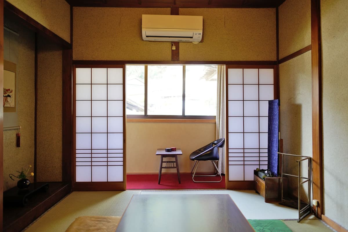 Ein traditioneller japanischer Raum mit Tatami-Böden, Shoji-Schirmen, einem Tatami-Tisch, einem schwarzen Stuhl und einem kleinen Tisch mit einem roten Gegenstand.