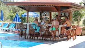 Un espace bar aménagé au bord de la piscine, avec des fauteuils en osier et un comptoir, le tout entouré de la piscine et de chaises longues.