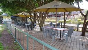 An outdoor patio with tables and chairs, a green railing, and trees providing shade. - Tents Hotel (Nairobi)