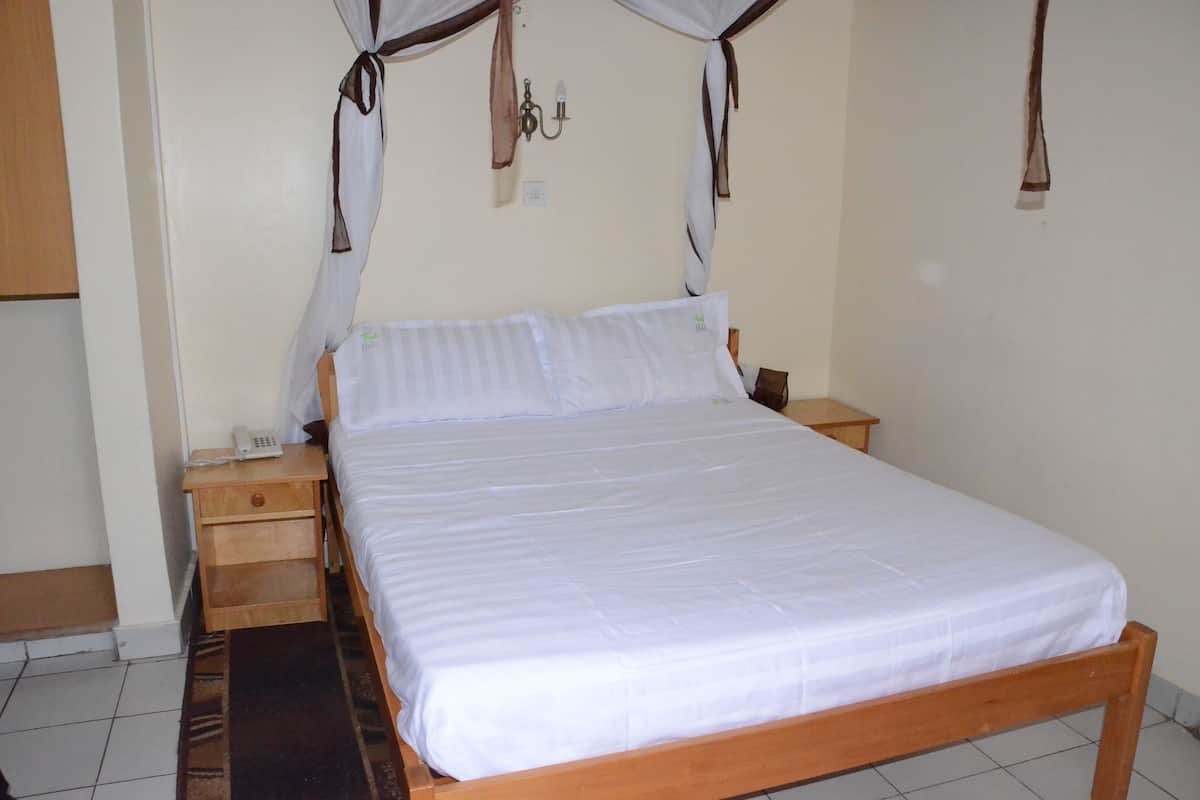 A single-bed room with a wooden frame, white bedding, a bedside table with a phone, and a wall-mounted light fixture.
