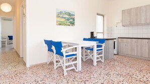 A kitchen with white cabinets, a white countertop, and a window. There is a painting on the wall and a white table with blue chairs. - Spacious Apartment on the Beachfront (Xylokastro-Evrostina)