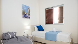 A bedroom with a bed, a bedside table with a lamp, a chair, a window with blinds, and a wall-mounted picture. - Spacious Apartment on the Beachfront (Xylokastro-Evrostina)