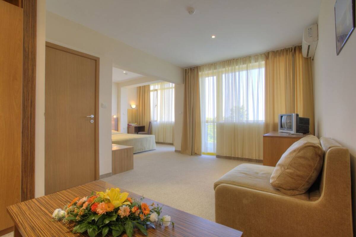 A hotel room with a wooden desk, a sofa, a television on a wooden side table, and a bed in the background.