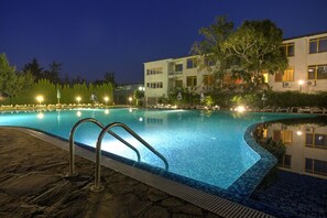 A swimming pool at night with a stone deck, surrounded by trees and a building with multiple windows and balconies.