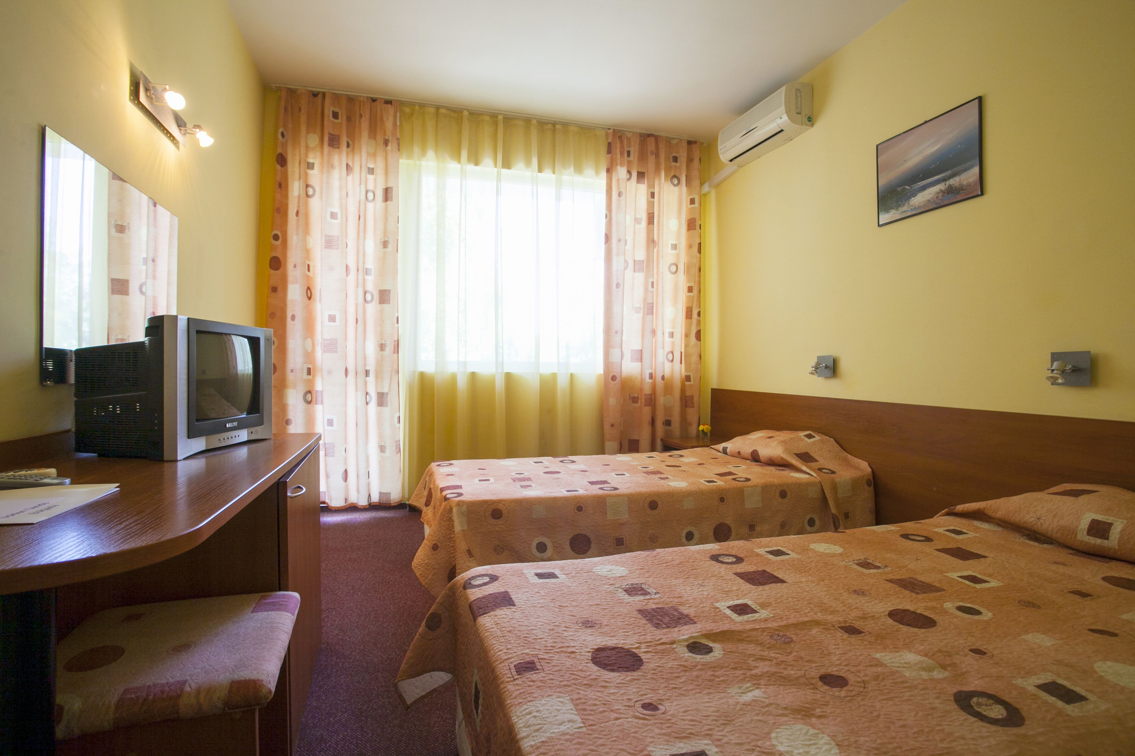 Habitación de hotel con dos camas, un escritorio con televisor, una ventana con cortinas y un aire acondicionado en la pared.