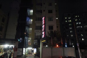 Un immeuble à plusieurs étages, doté d’une enseigne lumineuse, d’une entrée sécurisée par un portail et d’un magasin situé au niveau de la rue.