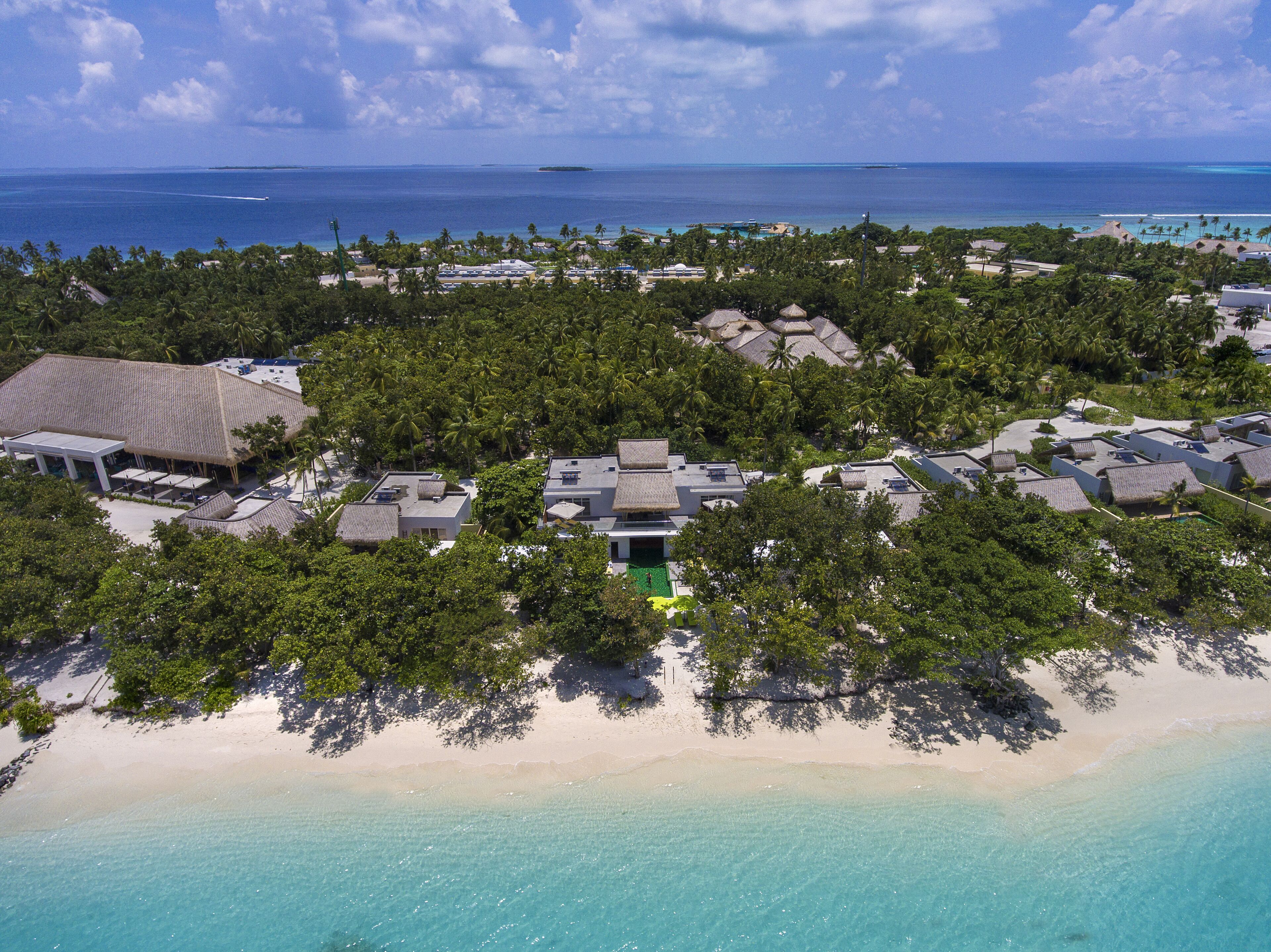 An aerial view of a resort with multiple bungalow-style buildings surrounded by dense tropical vegetation, a beach, and a clear view of the ocean.