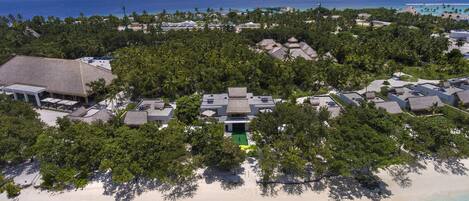 An aerial view of a resort with multiple bungalow-style buildings surrounded by dense tropical vegetation, a beach, and a clear view of the ocean.