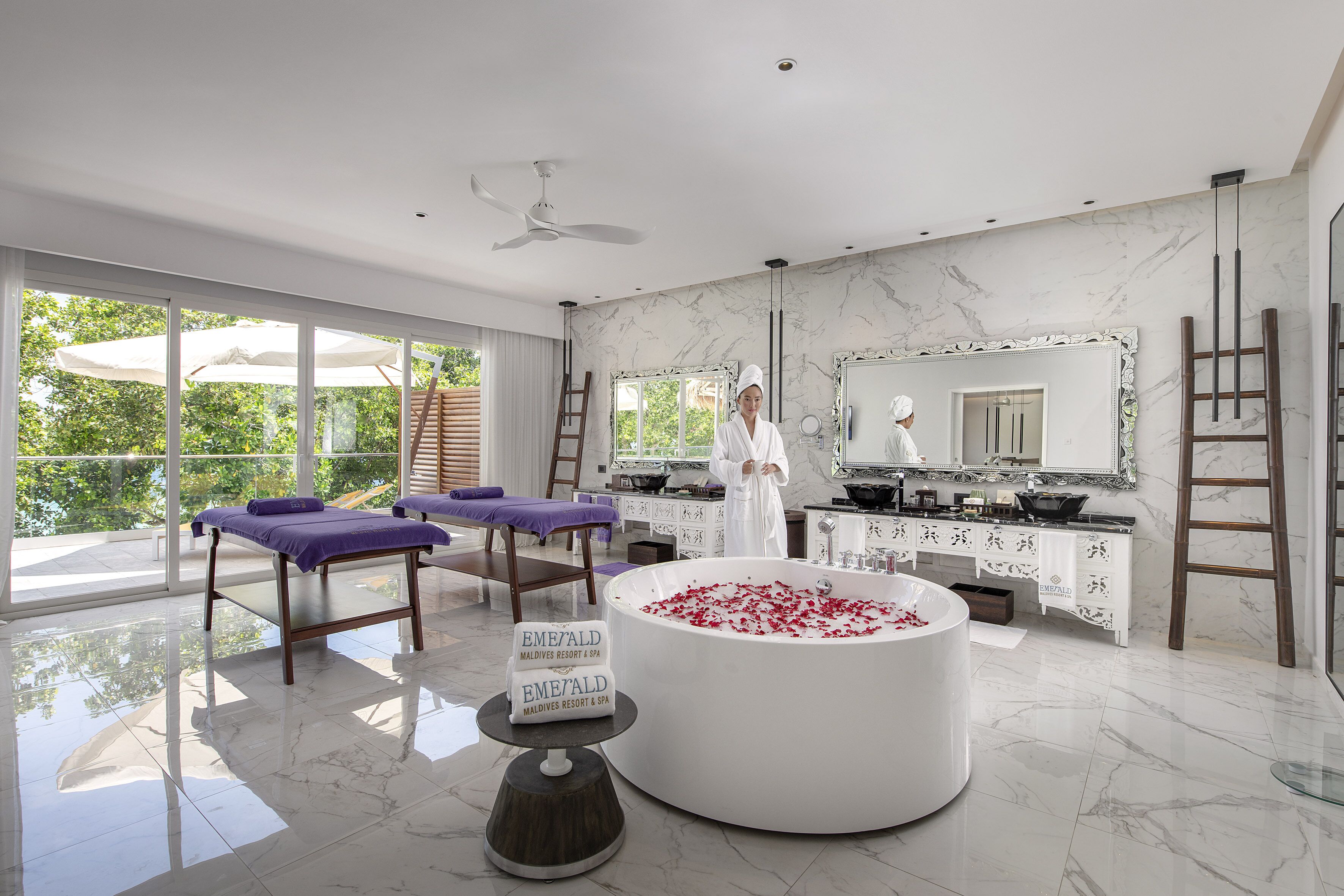 A spa room with a large bathtub filled with rose petals, a massage table, and a sign indicating the location as 'Emerald Maldives Resort & Spa'.