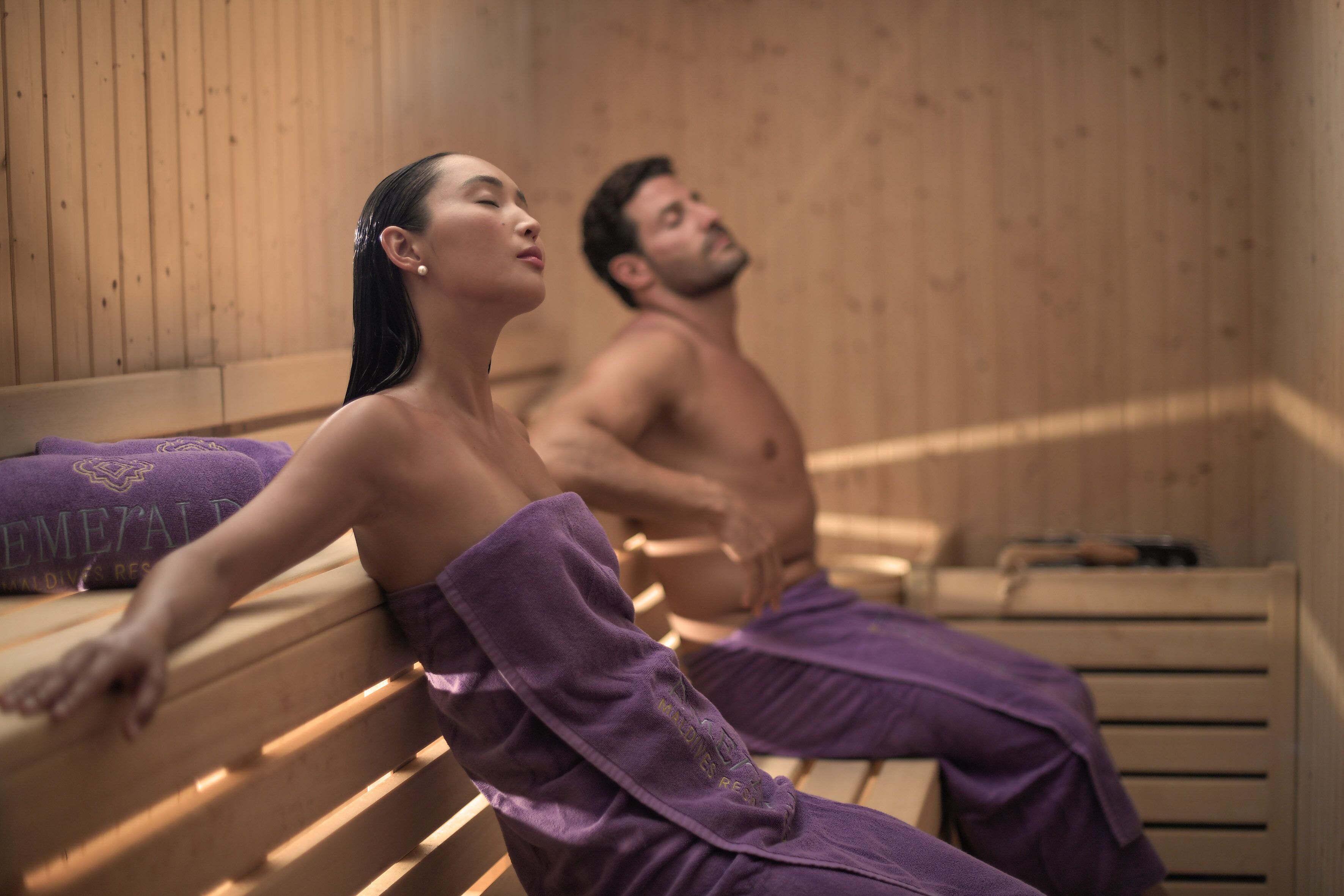 Two people in a sauna with wooden benches and purple towels.