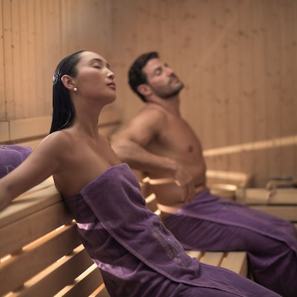 Two people in a sauna with wooden benches and purple towels.