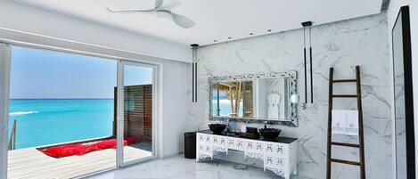 A modern bathroom with a large mirror, white marble flooring, and a view of the ocean.