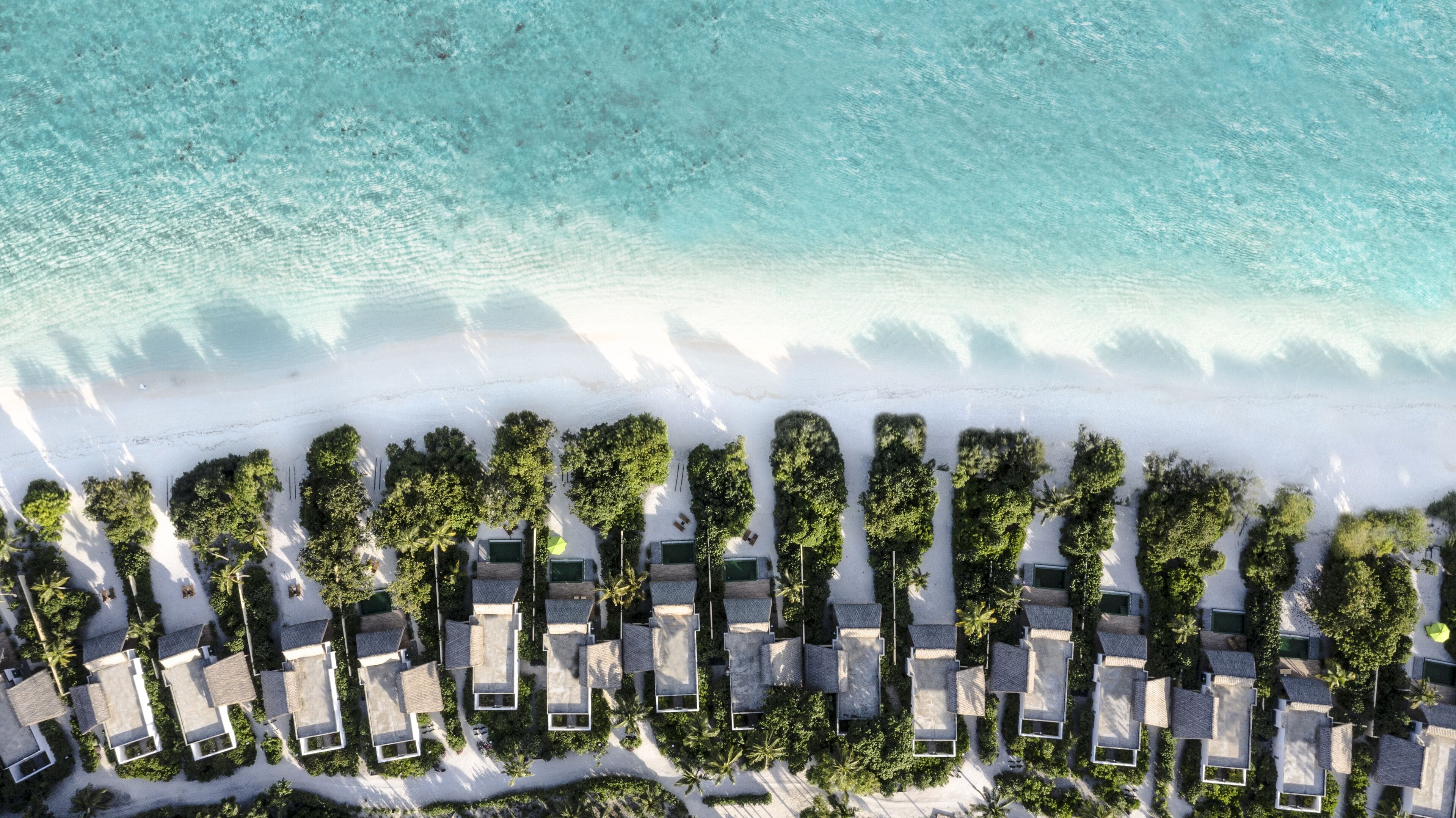 Vista aérea de un resort con hileras de alojamientos tipo bungaló alineados frente a una playa inmaculada.