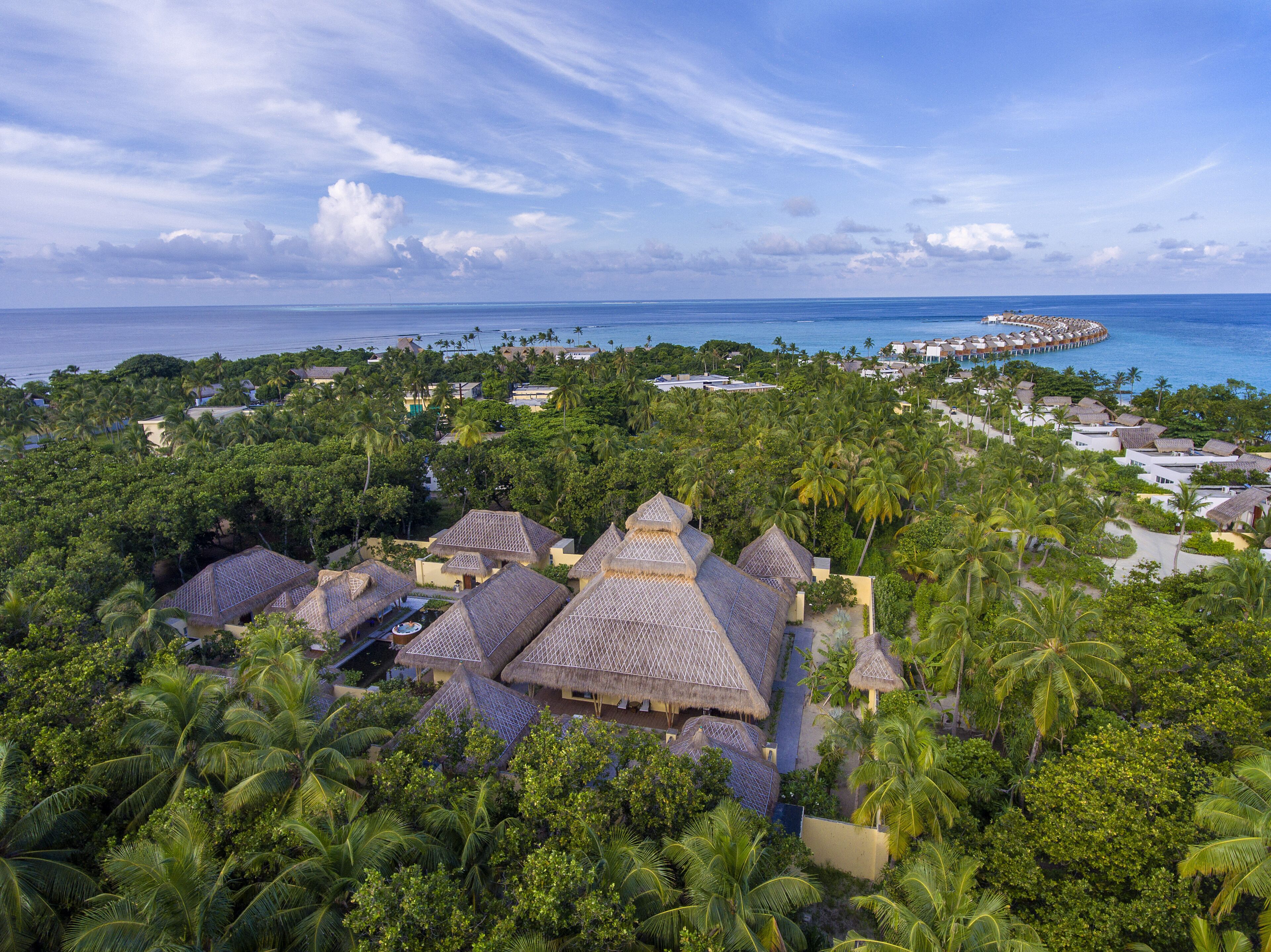 An aerial view of a resort with thatched-roof buildings surrounded by dense palm trees, a clear blue sky, and a distant overwater structure.