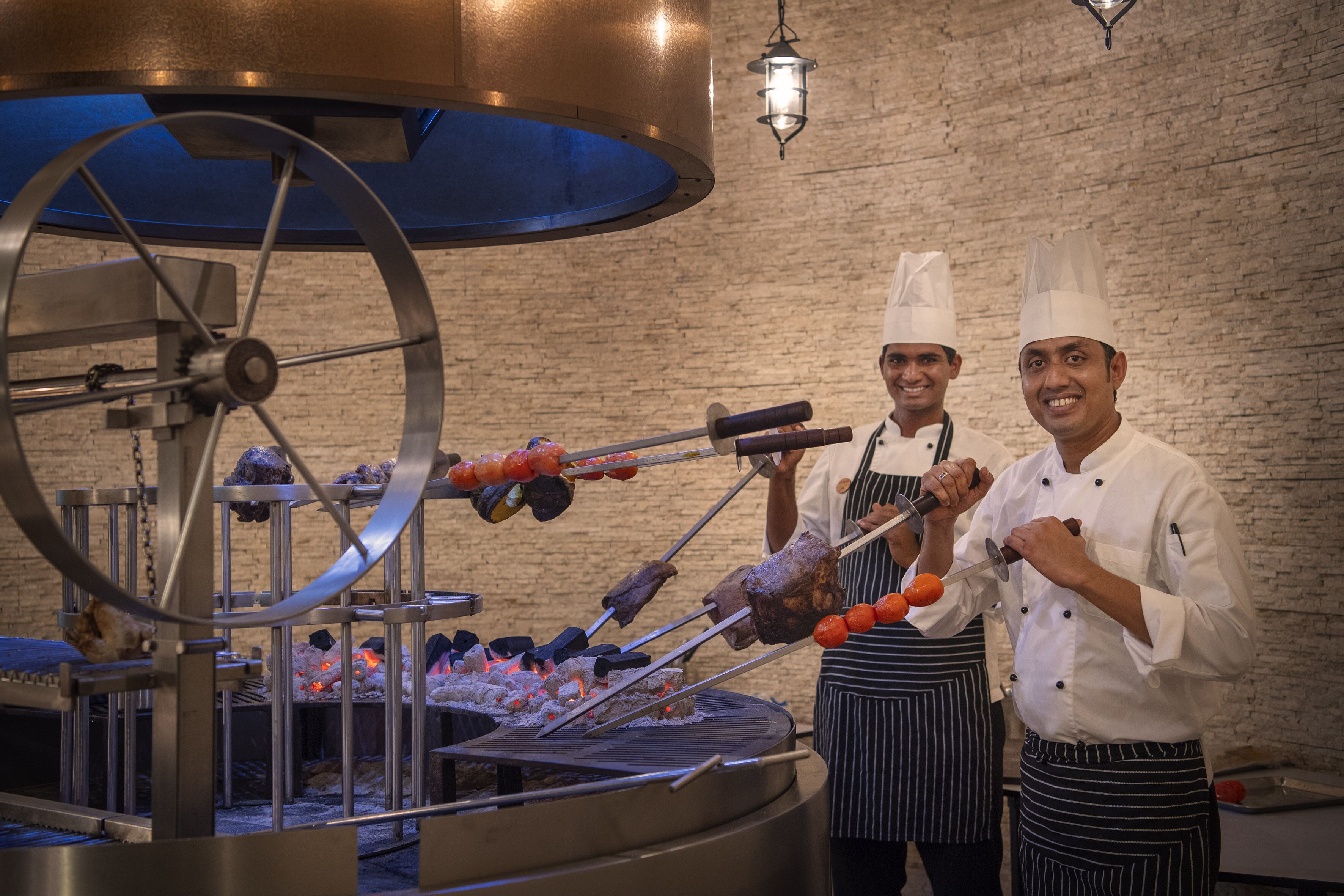 Cocineros preparando comida en una parrilla giratoria, en una cocina con pared de ladrillo de fondo.