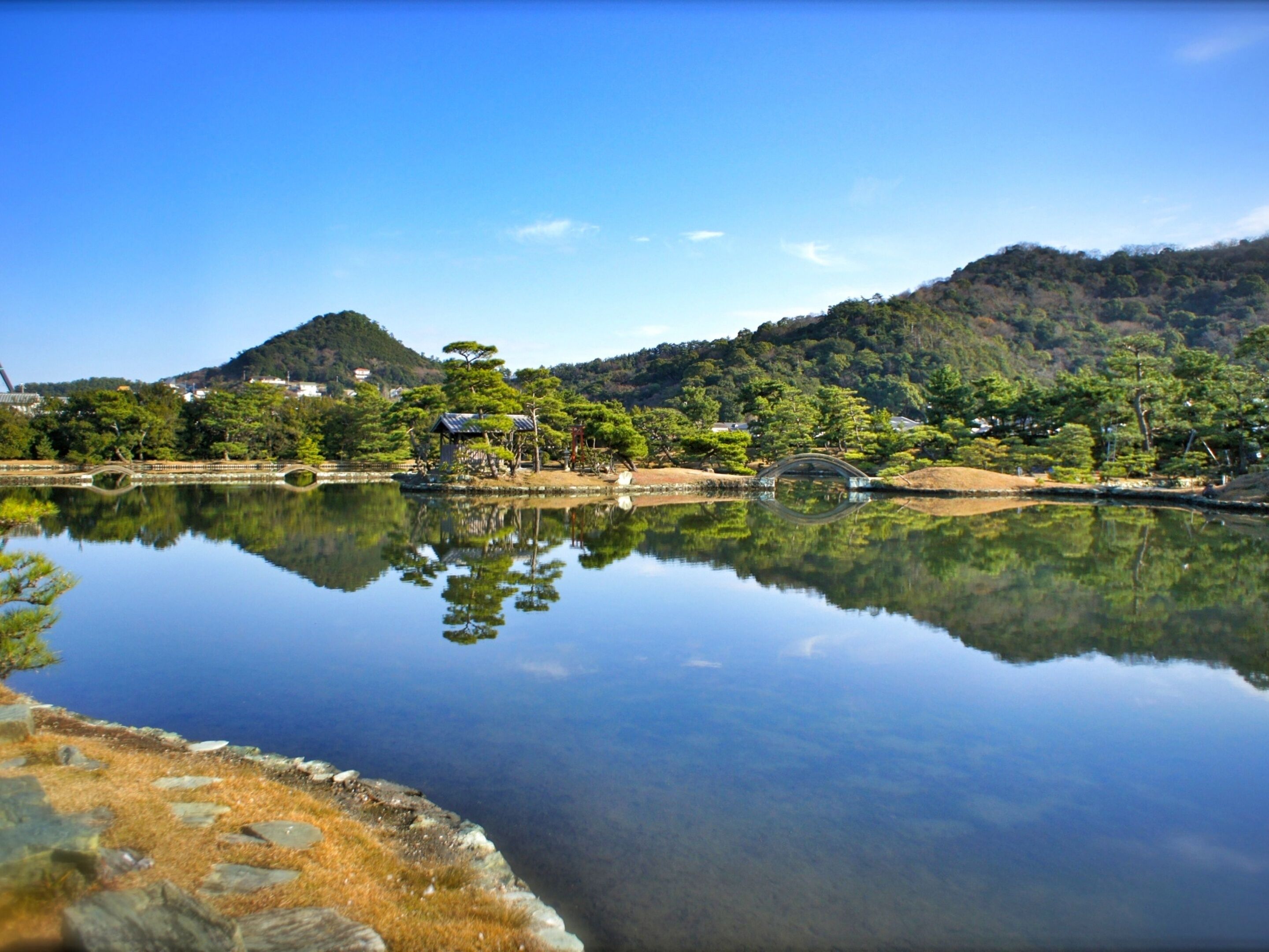 A tranquil lakeside setting with a bridge, trees, and a hill in the background.