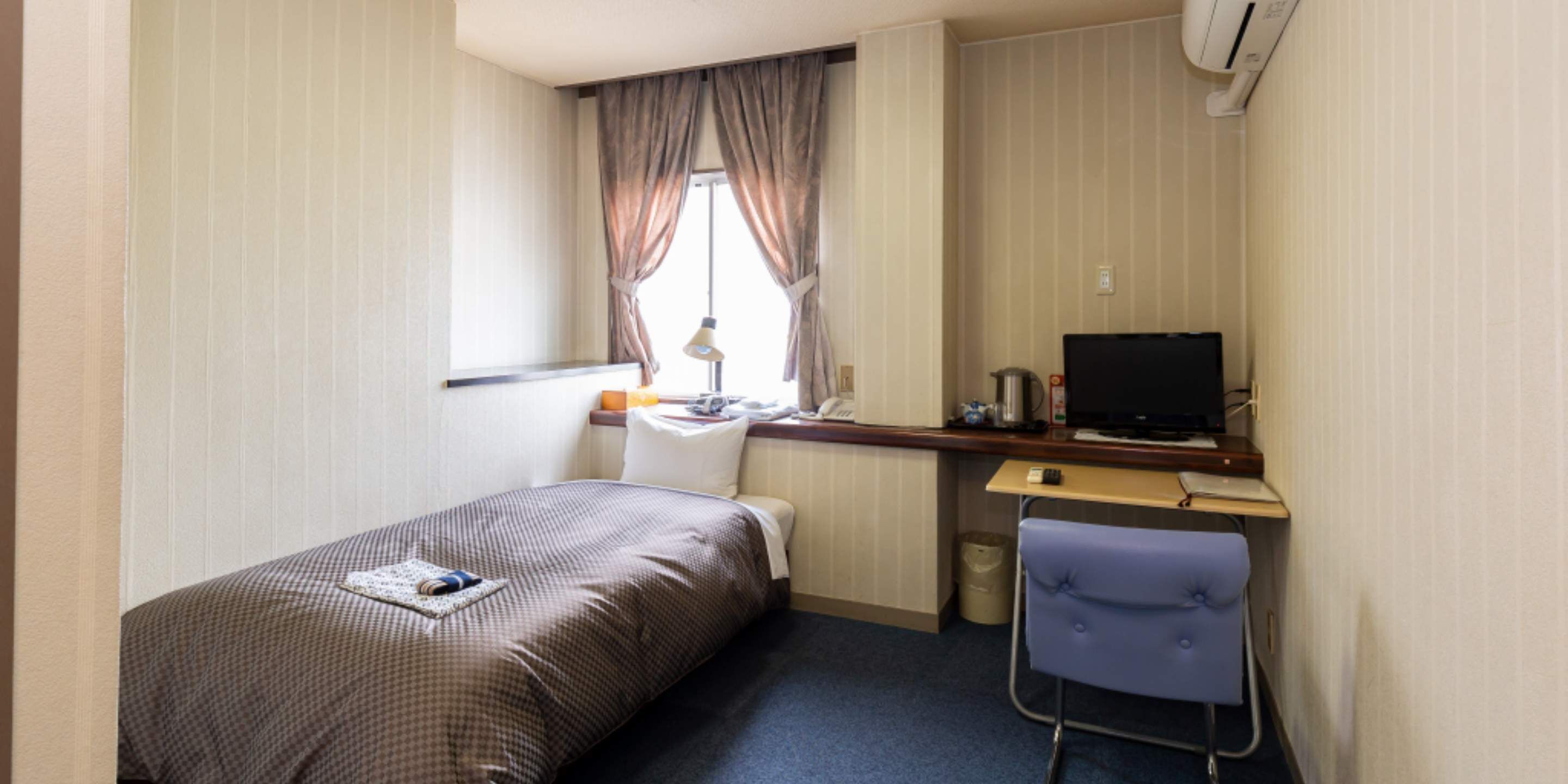 Una habitación de hotel pequeña y ordenada con una cama individual, un escritorio con ordenador, una silla y una ventana con cortinas.