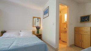 Un dormitorio con cama, mesita de noche, espejo, un cuadro, y un baño visible a través de una puerta abierta, además de un armario de madera.