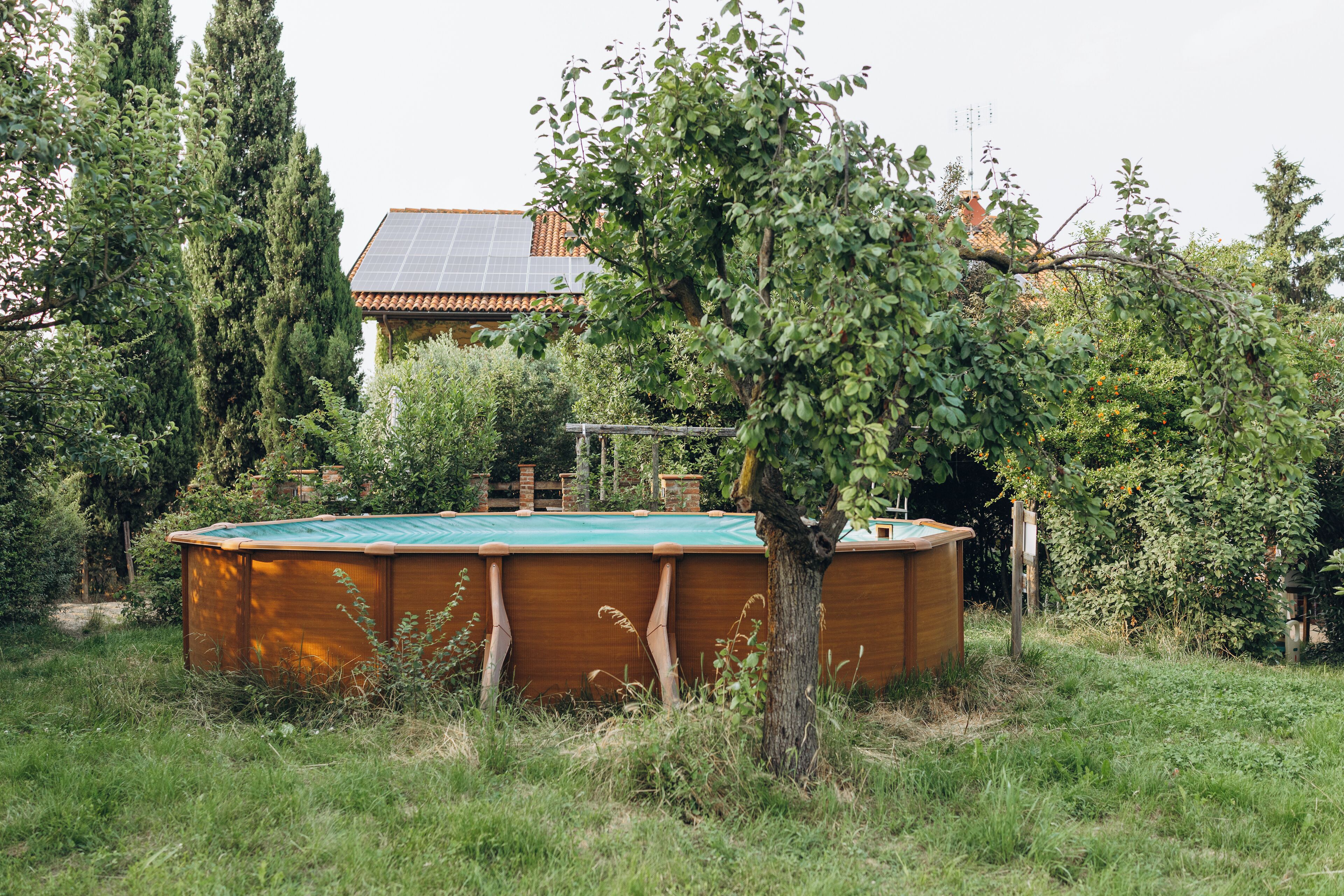 Una piscina de madera con una cubierta verde, rodeada de césped y árboles, con una casa y paneles solares al fondo.