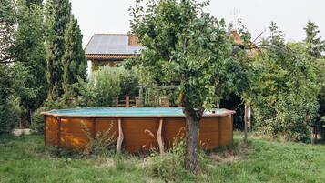 Una piscina de madera con una cubierta verde, rodeada de césped y árboles, con una casa y paneles solares al fondo.
