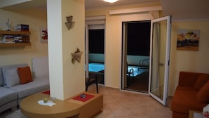 Una sala de estar con un sofá, una mesa de centro y una silla. Hay una estantería con libros y objetos decorativos. Una puerta corrediza de cristal da a un balcón con piscina.