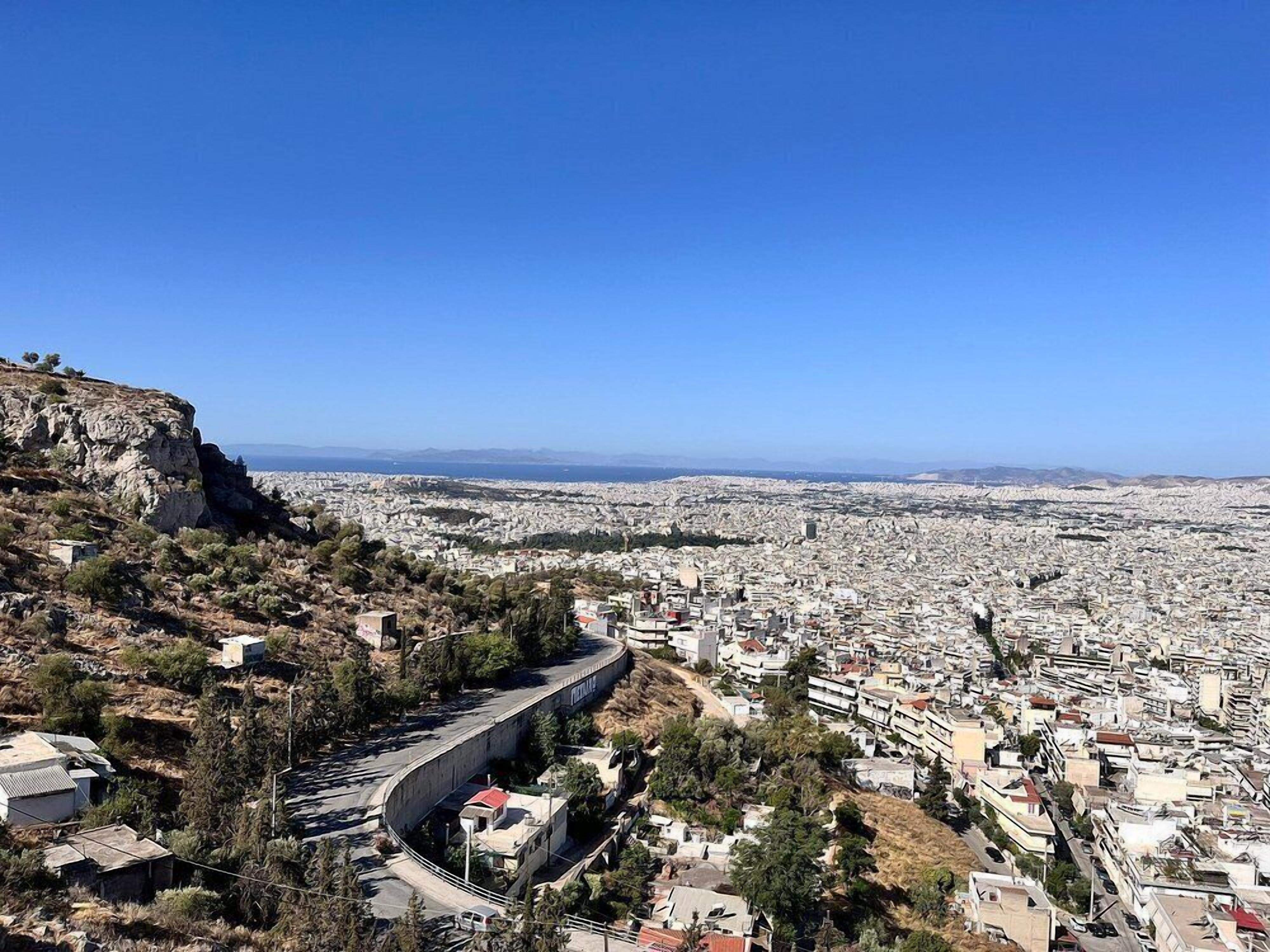 Un paysage urbain avec une route sinueuse bien visible sur une colline, menant à une zone urbaine densément peuplée, avec des bâtiments et un ciel bleu dégagé.