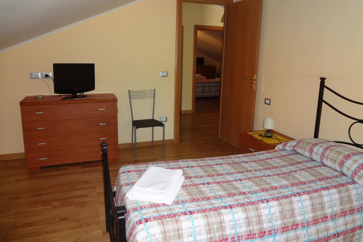 Een slaapkamer met een bed, een houten dressoir, een stoel, een televisie op het dressoir en een deur naar een andere kamer.