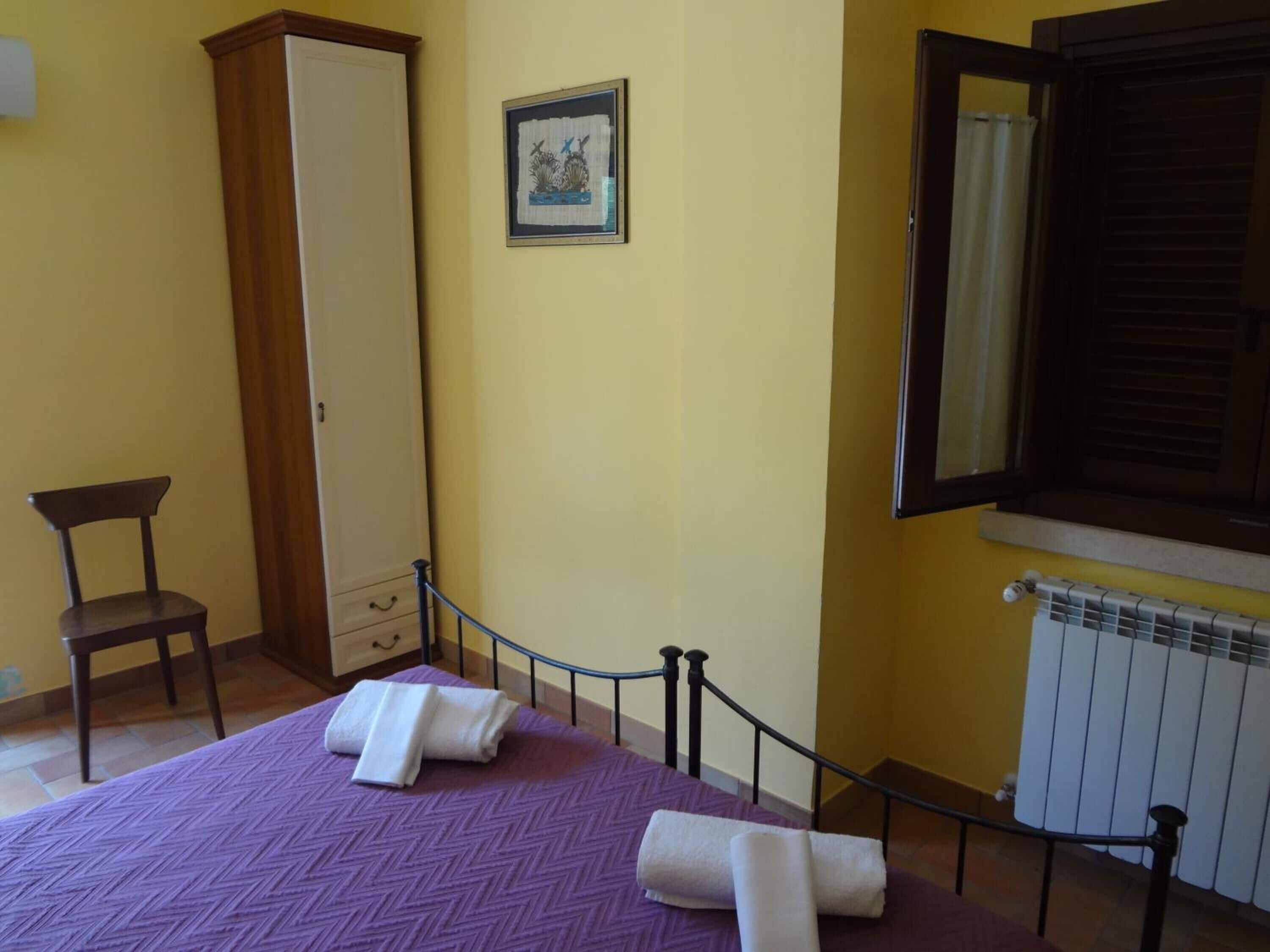 A bedroom with a purple bedspread, a wooden wardrobe, a framed picture on the wall, and a window with a curtain.