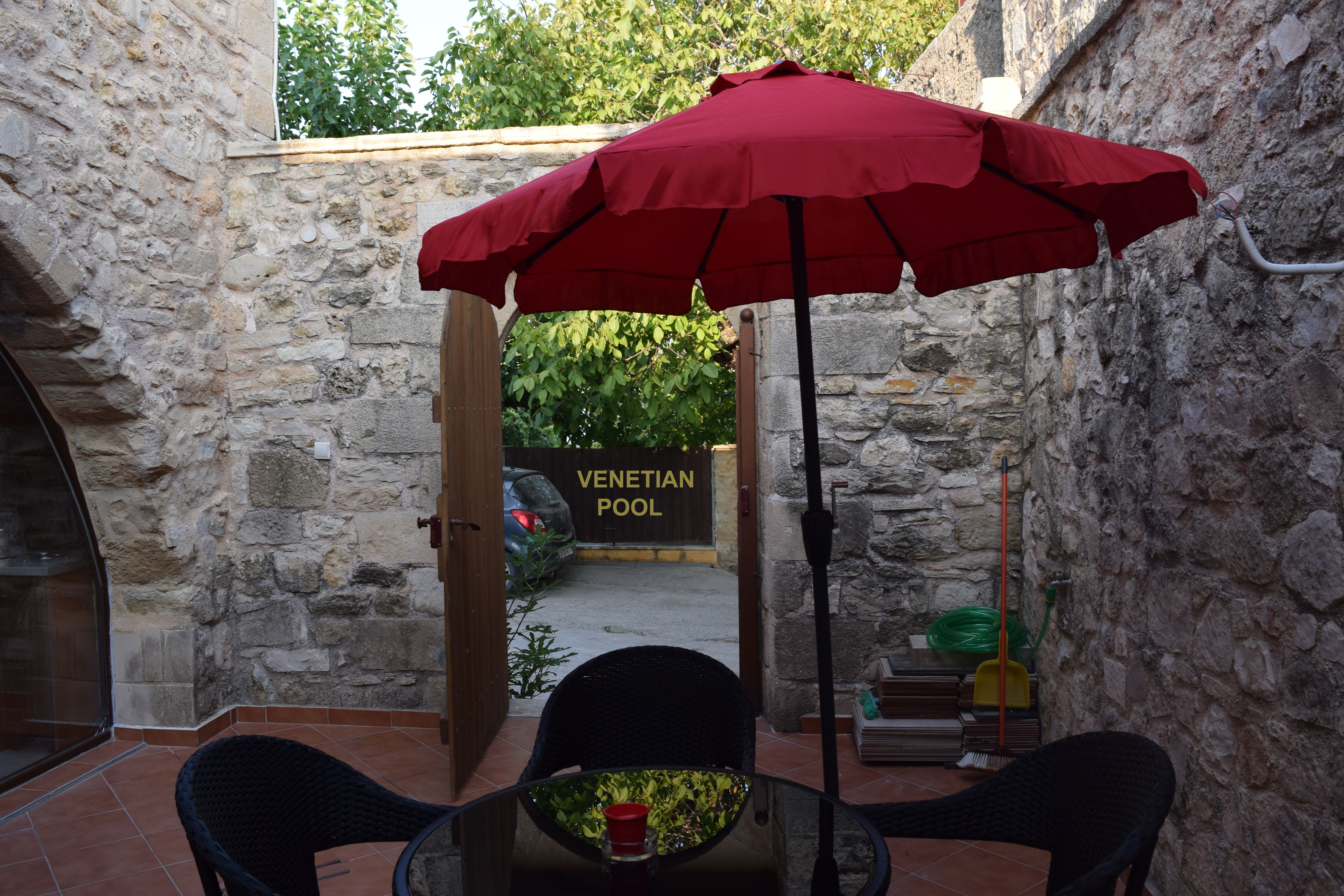 A stone-walled patio with a red umbrella, a table, and chairs. A sign for the Venetian Pool is visible in the background.