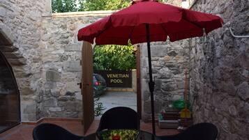 A stone-walled patio with a red umbrella, a table, and chairs. A sign for the Venetian Pool is visible in the background.