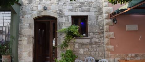 A stone building with a wooden door, a small window, and a potted plant. There is a table and chairs outside.