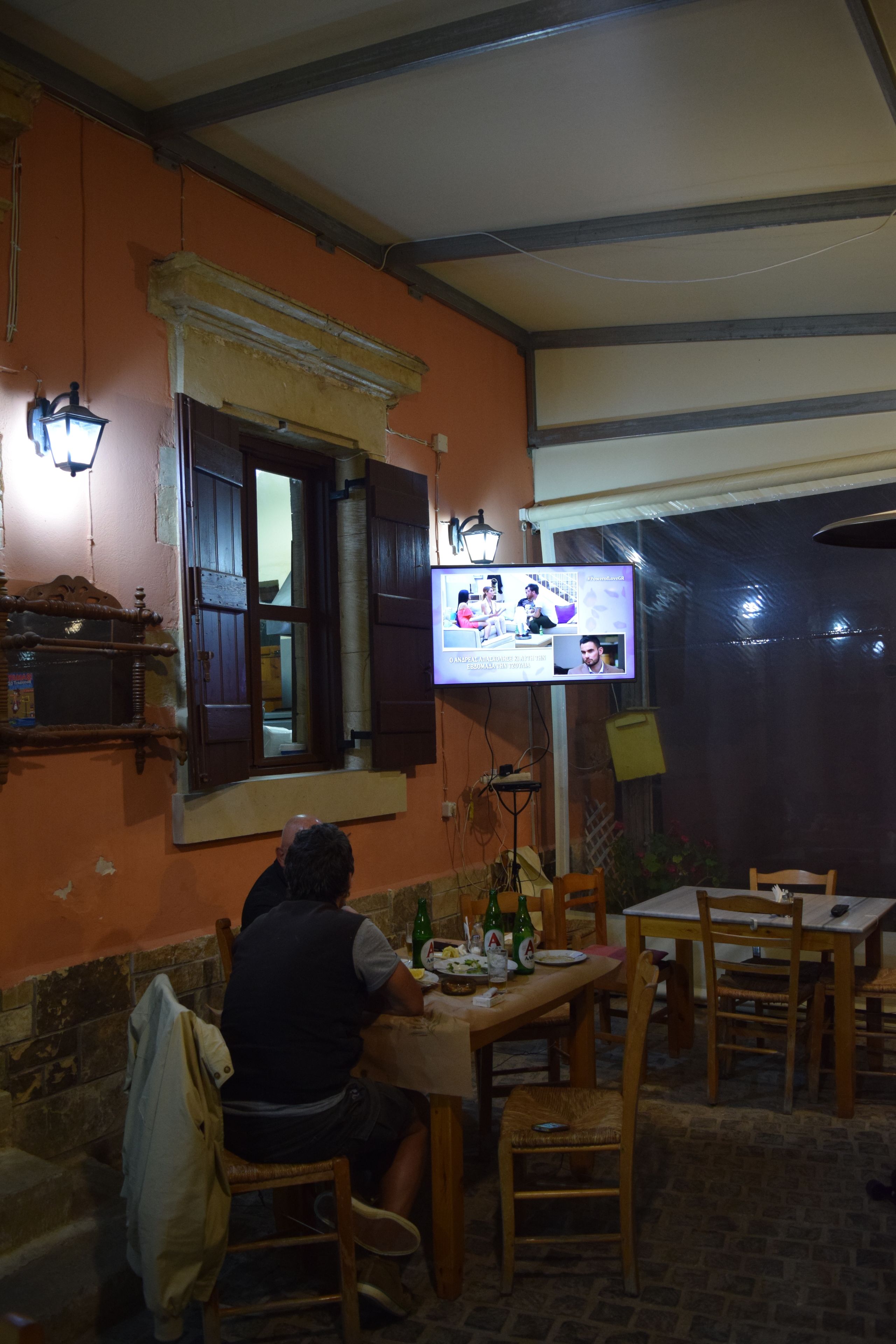 A person sitting at a table with a television displaying a program. The room has wooden furniture, a window with shutters, and a brick wall.
