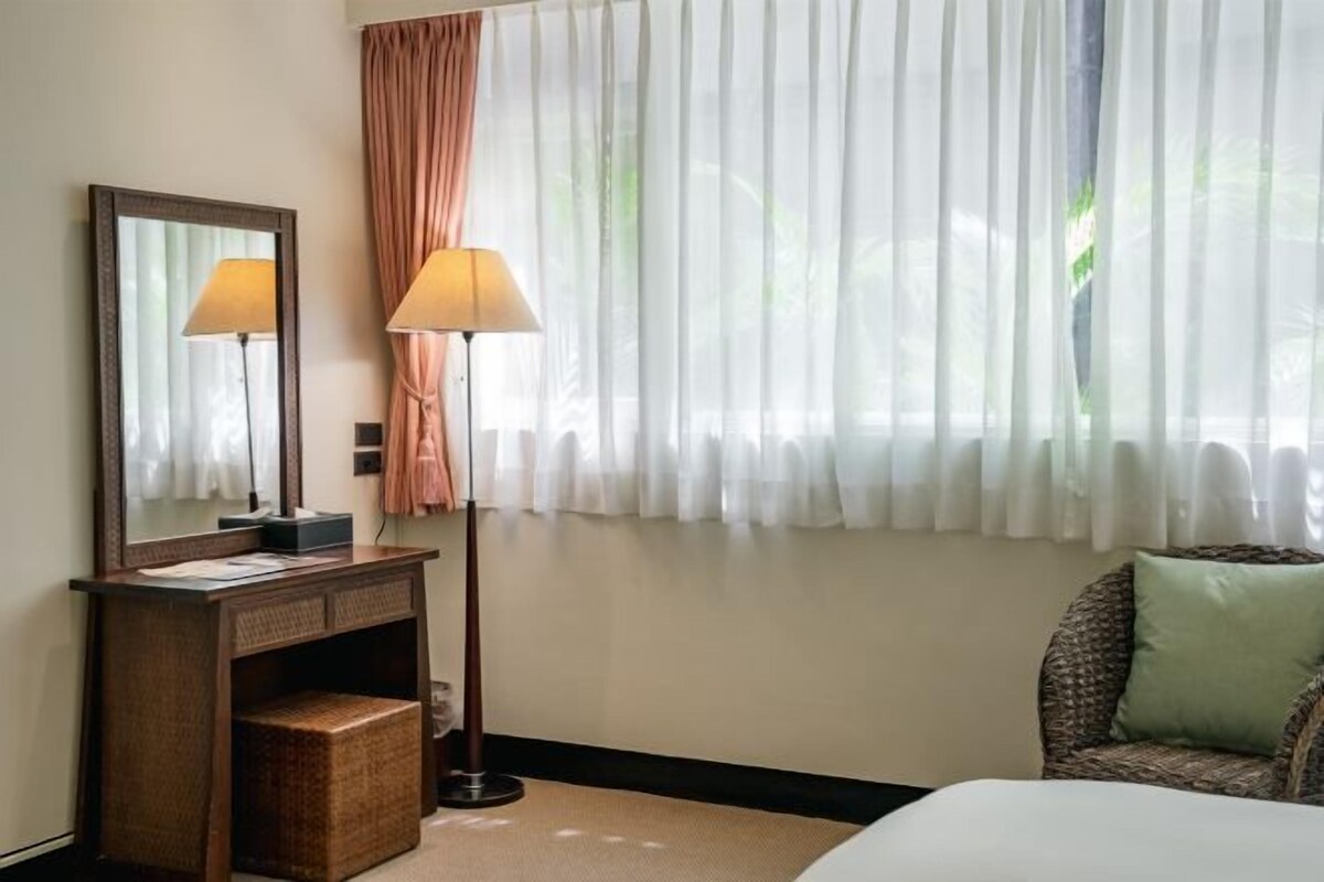 A hotel room with a bed, a bedside table with a lamp, a mirror, and a window with sheer curtains.