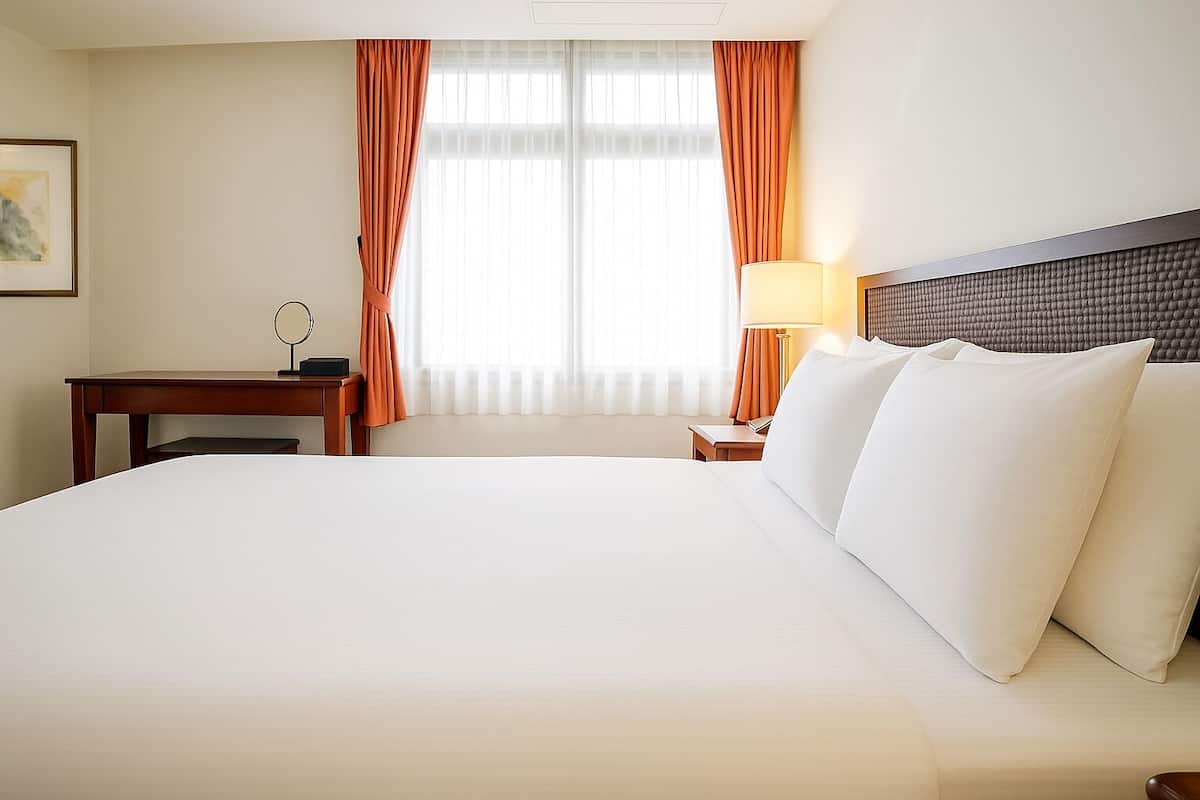 Quarto de hotel com uma cama grande, uma escrivaninha, uma luminária e uma janela com cortinas.