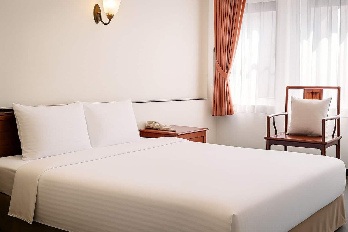 Quarto de hotel com uma cama grande, uma mesinha de cabeceira de madeira, uma cadeira e luminária fixada na parede.