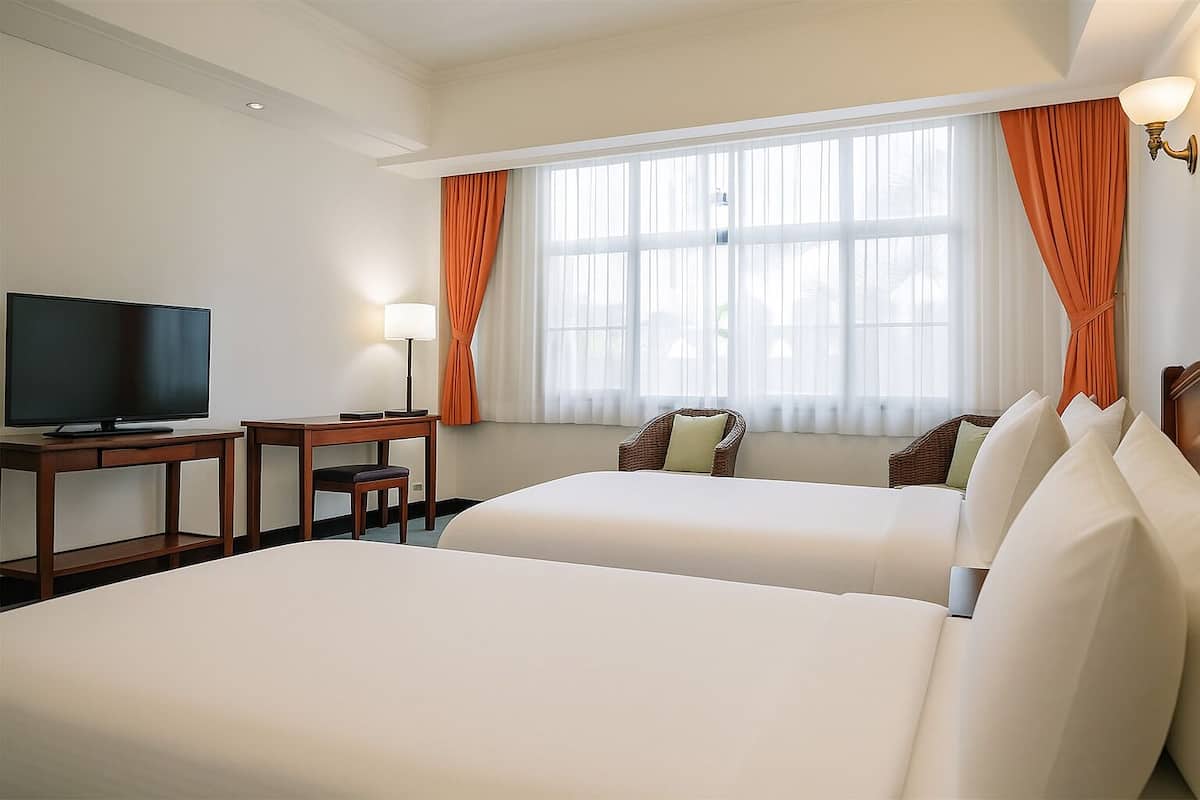 Quarto de hotel com duas camas, uma TV, uma mesa de trabalho com cadeira e uma janela com cortinas.