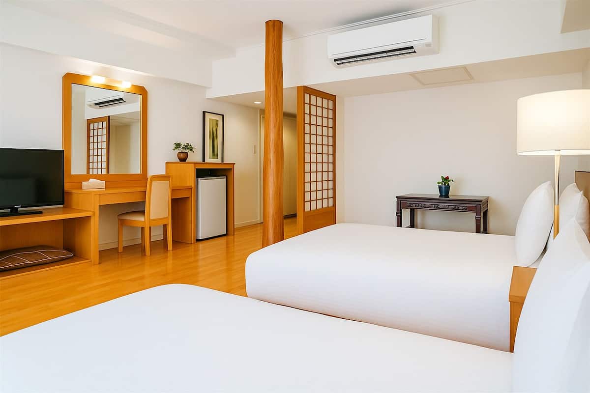 Quarto de hotel com uma cama, uma escrivaninha com cadeira, televisão, uma mesinha de cabeceira com abajur e um aparelho de ar condicionado.