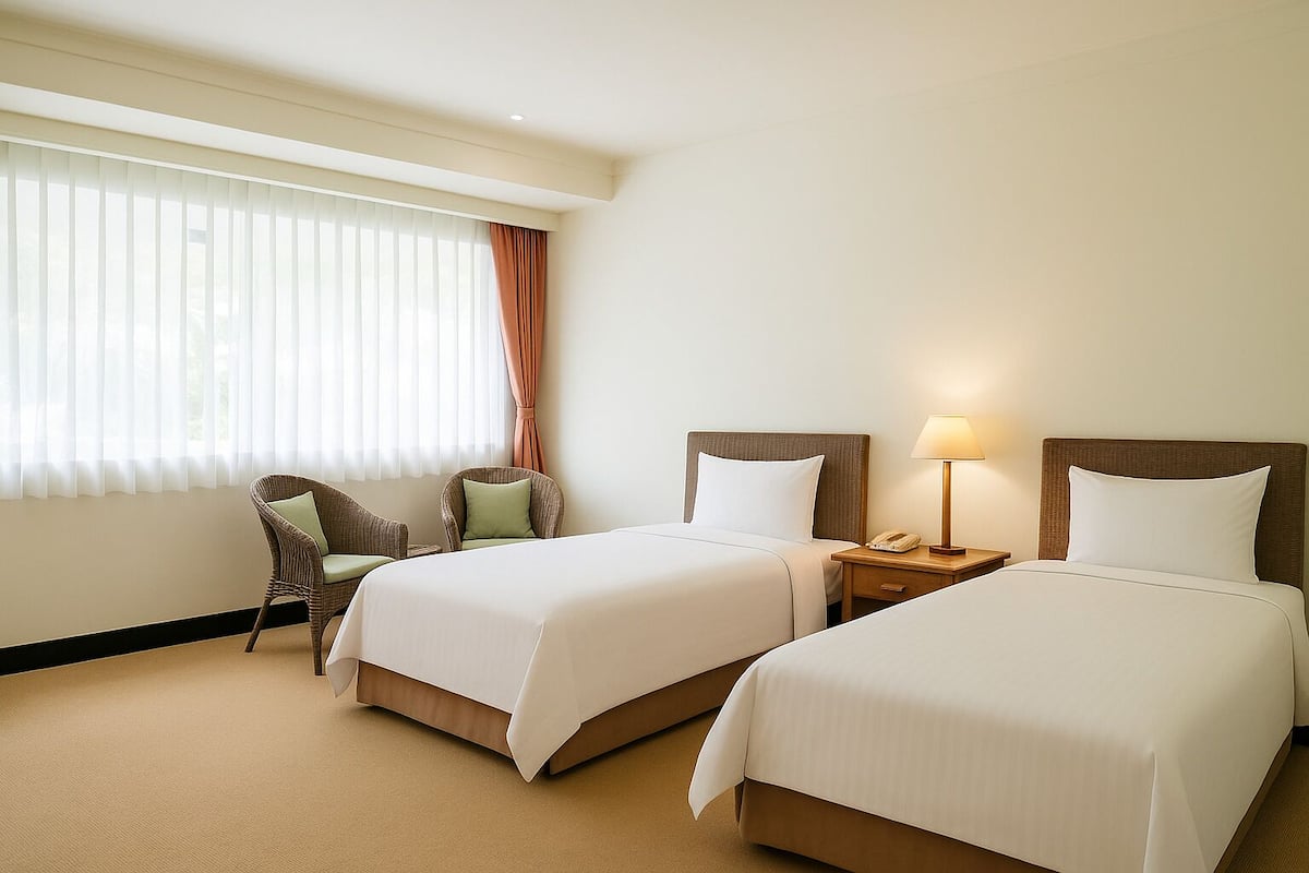 Quarto de hotel com duas camas, uma cadeira, uma mesa de cabeceira com abajur e uma janela com persianas.