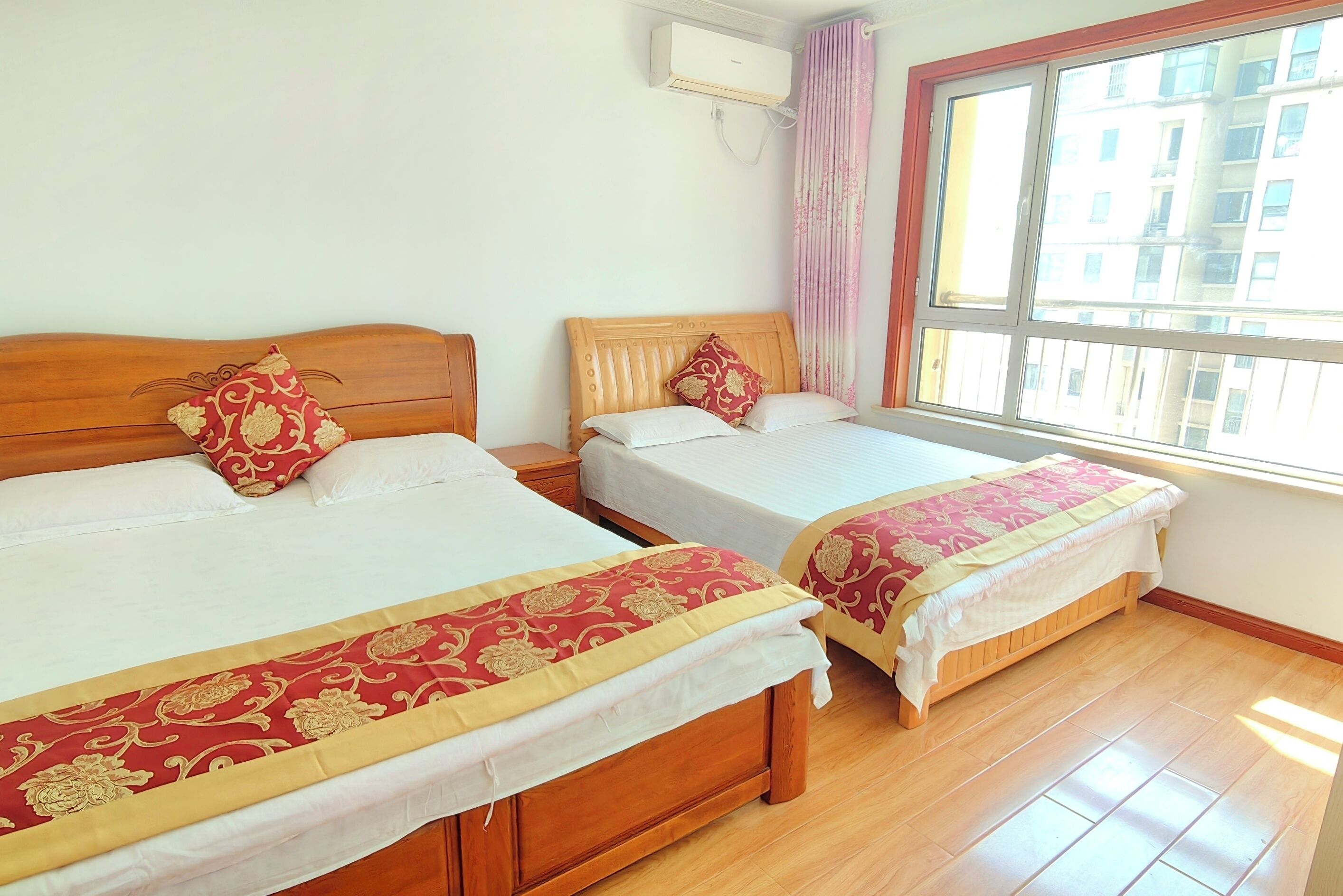 Two wooden beds with white linens and red decorative pillows, a window with a view of buildings, and an air conditioning unit on the wall.