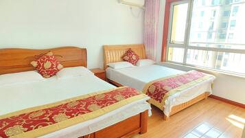 Two wooden beds with white linens and red decorative pillows, a window with a view of buildings, and an air conditioning unit on the wall.