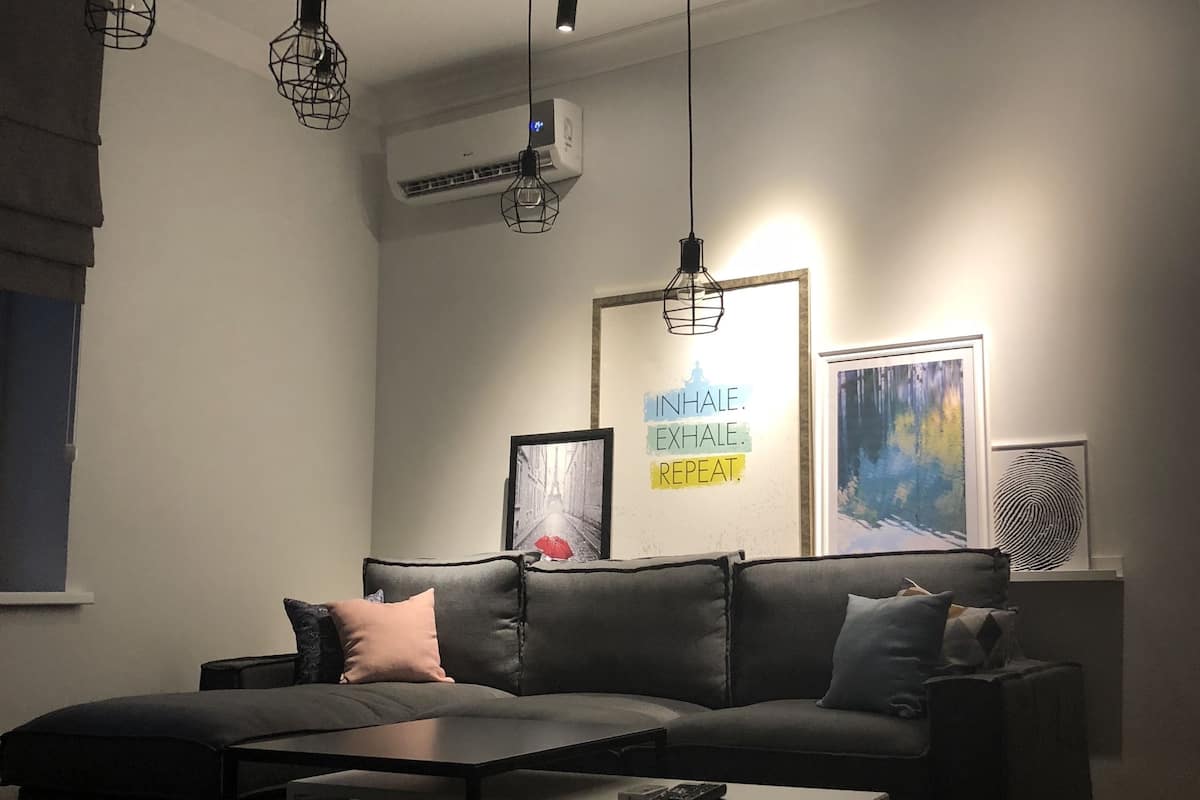 Una sala de estar moderna con un sofá seccional, una mesa de centro y lámparas colgantes. Hay un equipo de aire acondicionado en la pared y una obra de arte enmarcada sobre el sofá.