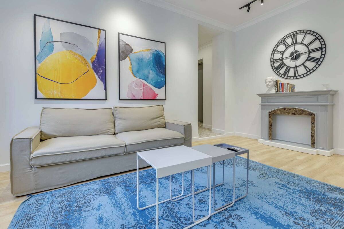 Un salón con un sofá gris, una mesa de centro, cuadros abstractos, un reloj y una chimenea.